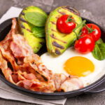 Grilled bacon and avocado, fried eggs with spinach and cherry tomatoes in cast-iron pan. Gray concrete background. Ketogenic diet. Low carb high fat breakfast. Healthy food concept