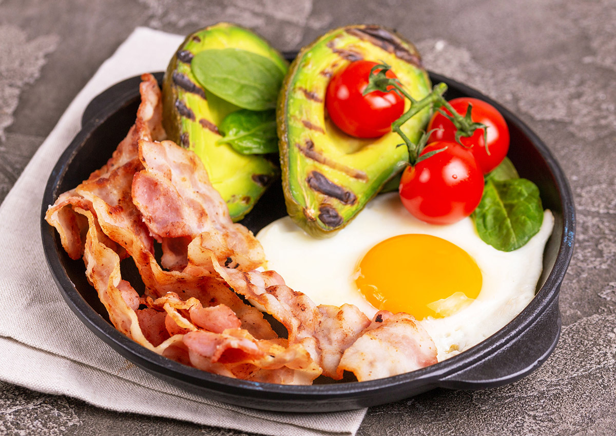 Grilled bacon and avocado, fried eggs with spinach and cherry tomatoes in cast-iron pan. Gray concrete background. Ketogenic diet. Low carb high fat breakfast. Healthy food concept